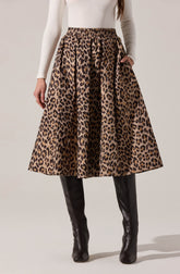 A woman is wearing a high-waisted, full-length skirt with a bold leopard print pattern in shades of brown and black. She pairs it with black, knee-high leather boots and a fitted, long-sleeved white top.